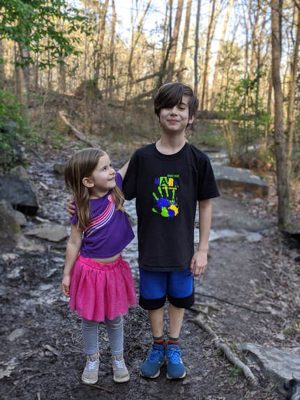 Juniper in a purple shirt and pink skirt with her arm around her brother, Cade, in a black tshirt and blue athletic shorts posing on a hiking trail