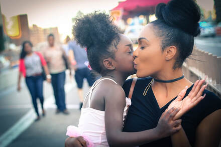 Photograph of black mother kissing her young daughter who is on her hip on the cheek 